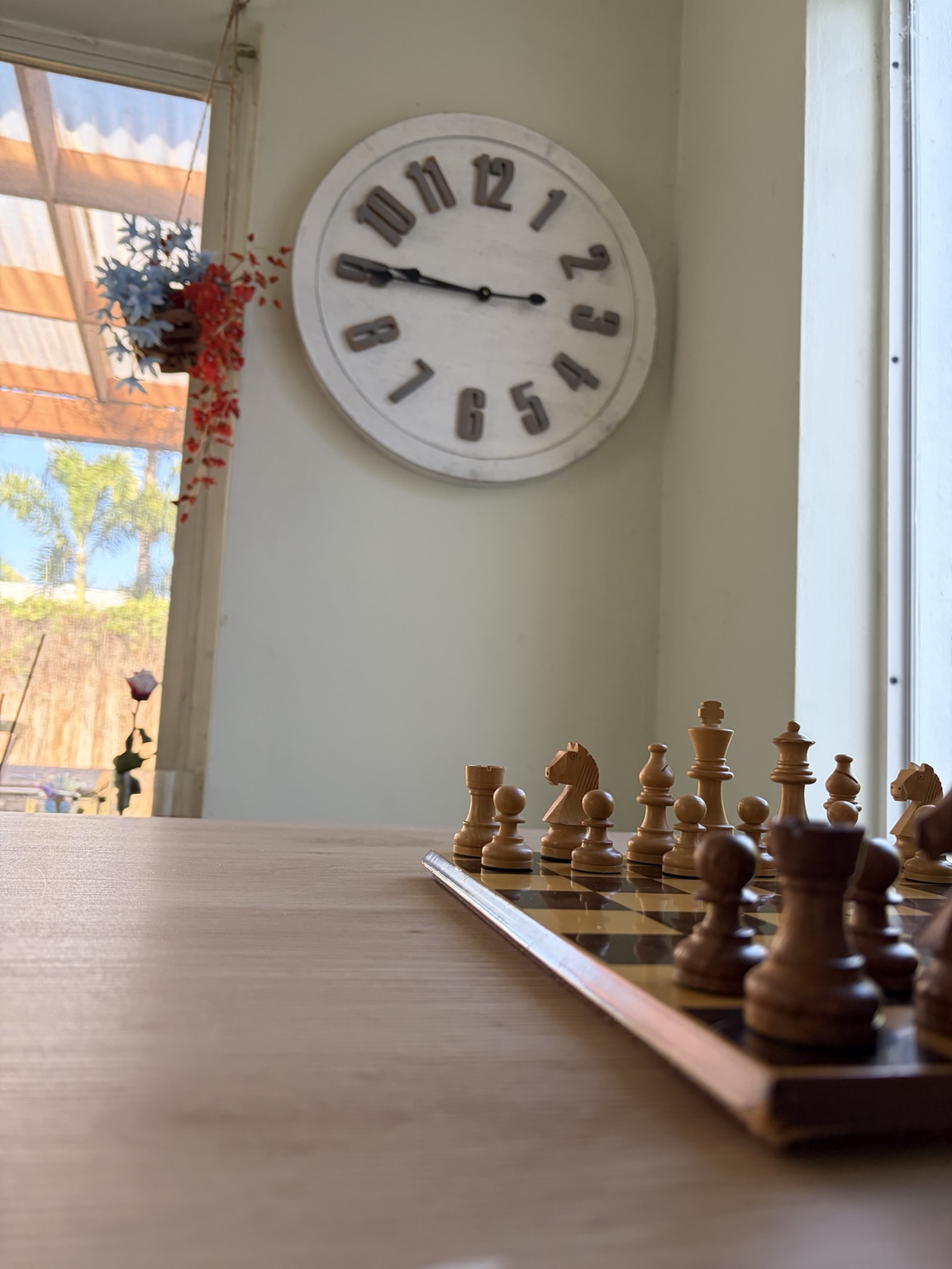 Wooden chess pieces on a board near a wall clock and sunlit window